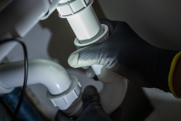 A person wearing black gloves adjusts a white PVC pipe fitting under a kitchen sink. The setting is dimly lit, highlighting the plumbing work being done in a home.