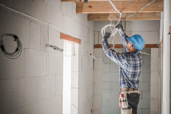Caucasian Electrician Installing Power Line in Newly Built Residential Unit Pulling Cables Under the Ceiling. Construction Site in the Background. Industrial Theme.
